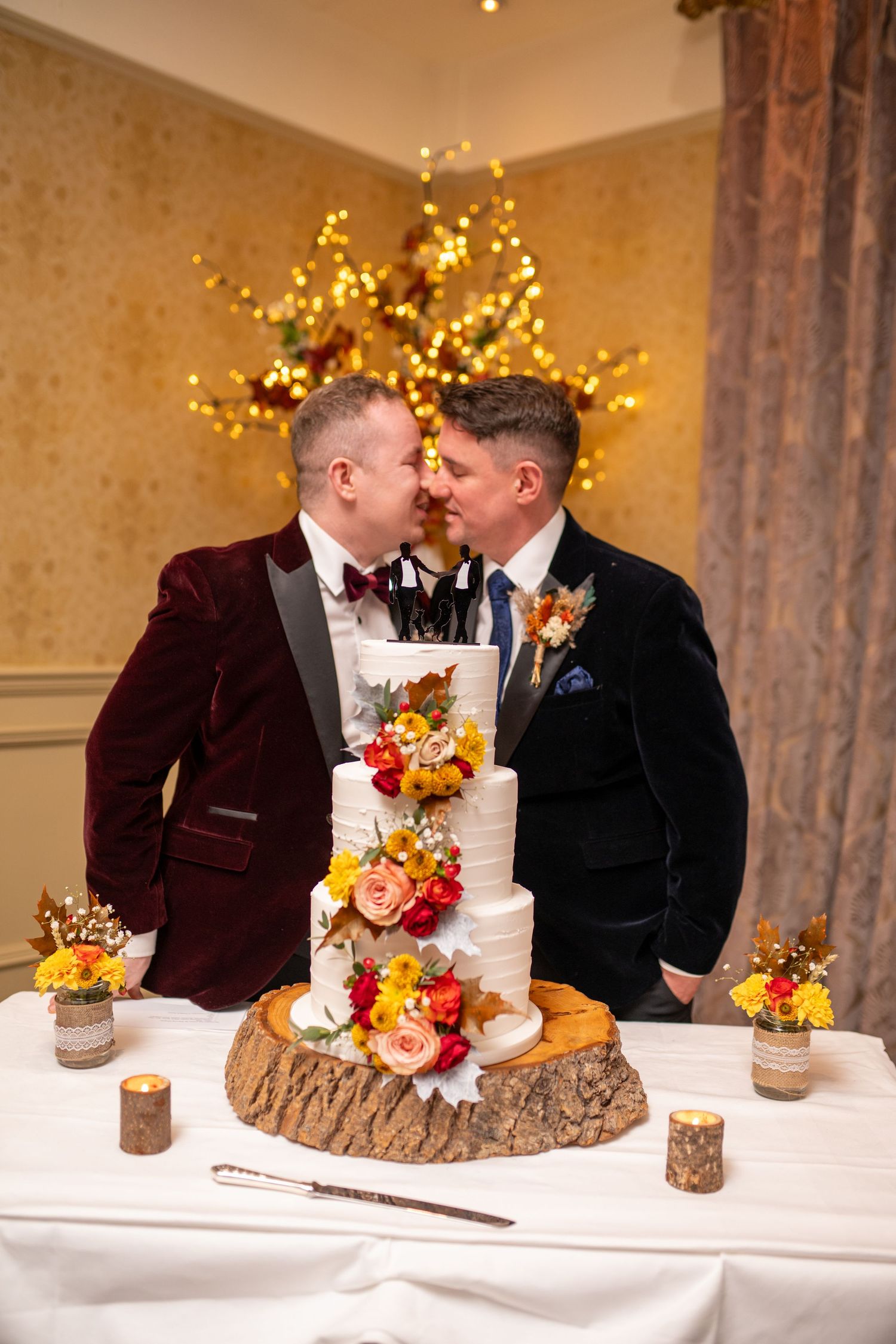 Two grooms share a kiss behind a floral wedding cake at their reception.