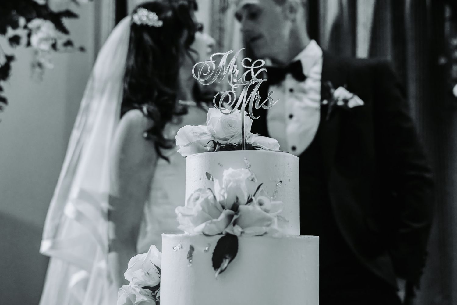 Black and white photo of a wedding cake with Mr & Mrs topper, adorned with roses, as bride and groom stand behind it.