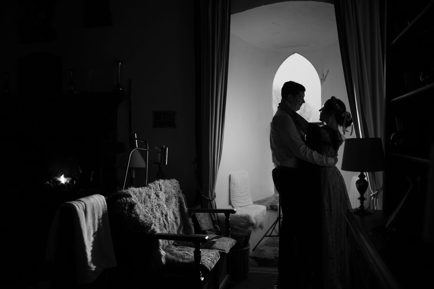 A romantic black and white silhouette of a couple embracing by an arched window indoors.