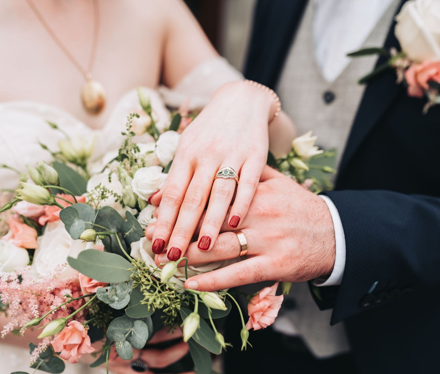 Newlyweds displaying wedding rings over a bridal bouquet of pink roses and eucalyptus, bride wearing red nail polish.