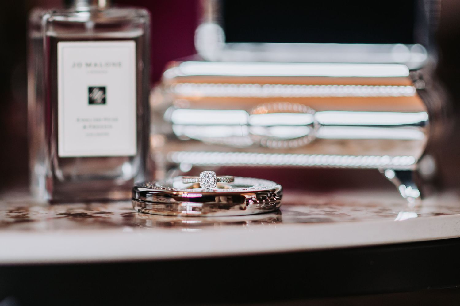 Wedding rings and diamond engagement ring displayed with Jo Malone perfume and jewelry box on marble surface.