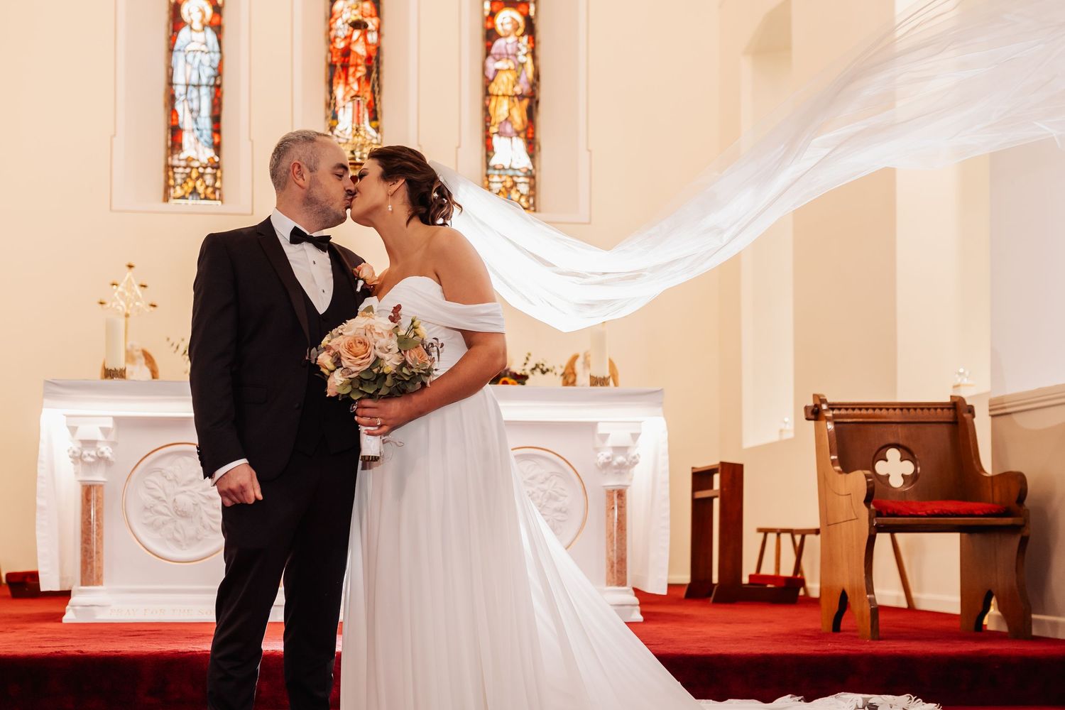 Bride and groom share a kiss at church altar with veil flowing dramatically in the air during wedding ceremony.