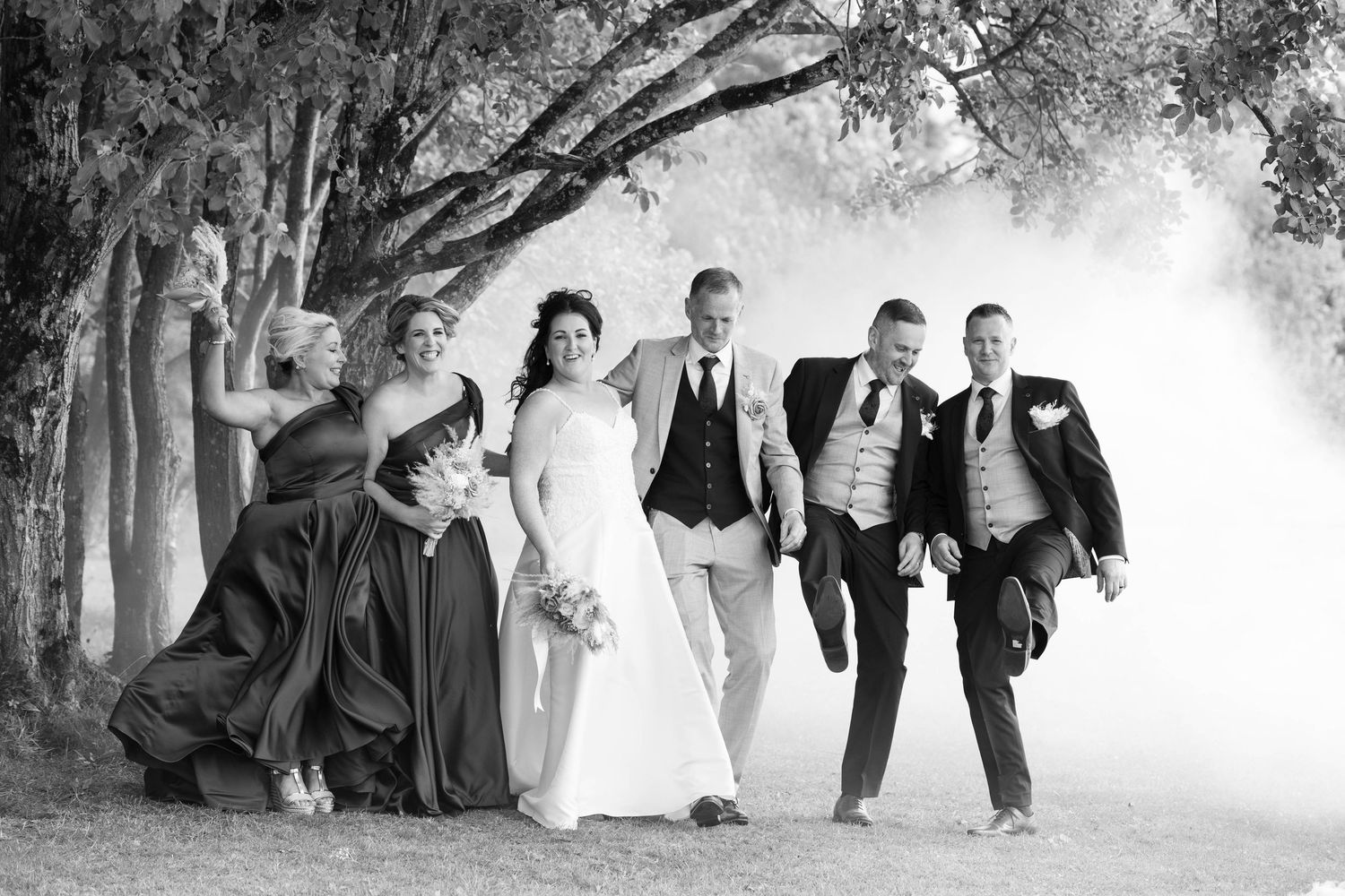 Black and white wedding party walking through dramatic smoke among trees.