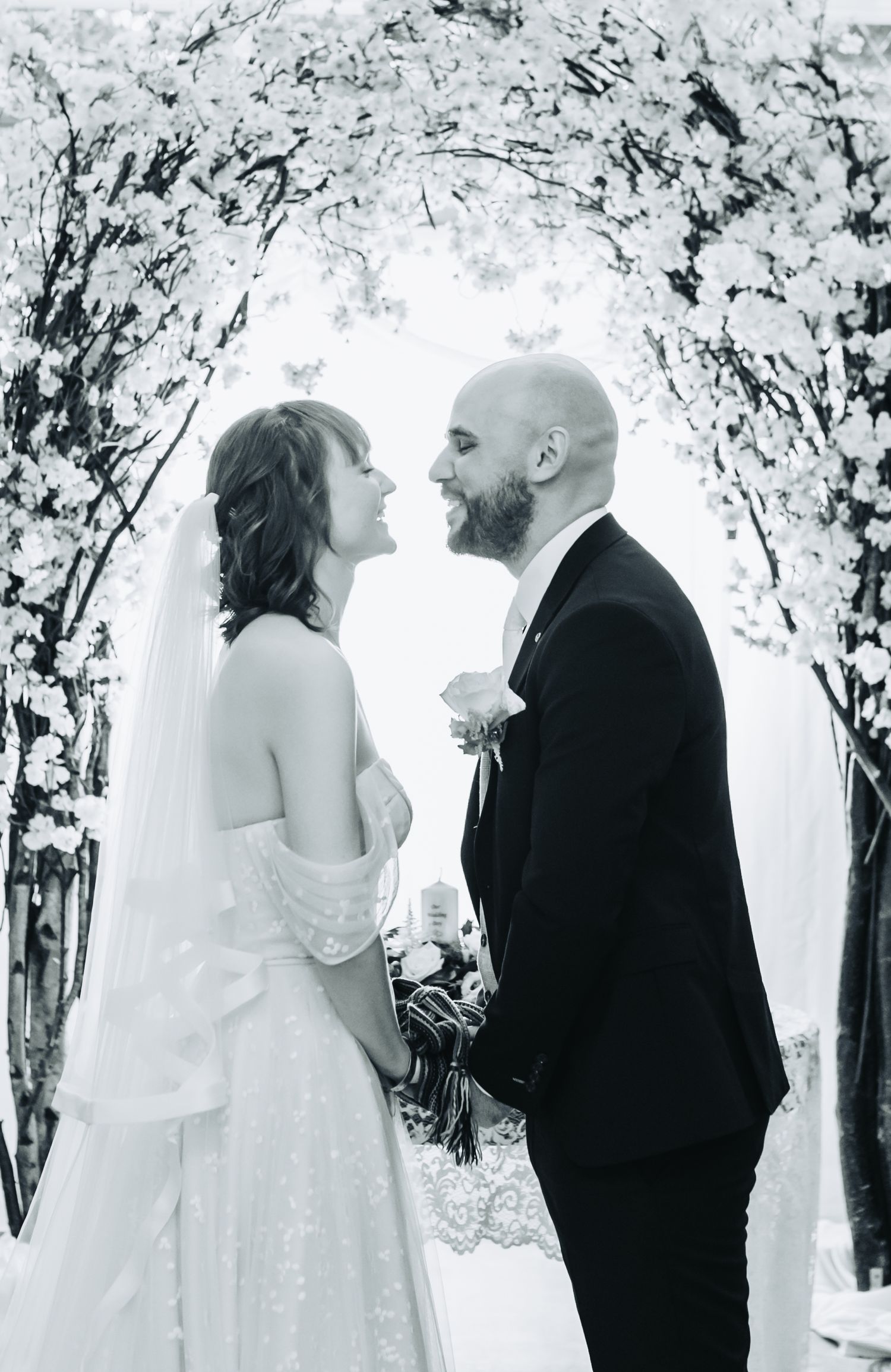 Black and white photo of bride and groom exchanging vows under a floral arch at their wedding ceremony.
