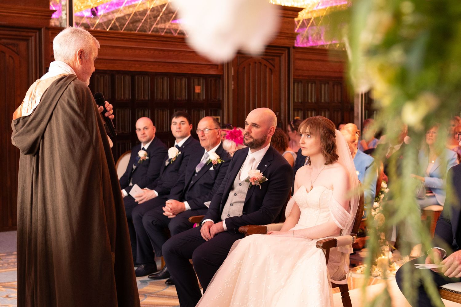 Bride and groom seated during wedding ceremony while officiant speaks at elegant venue.