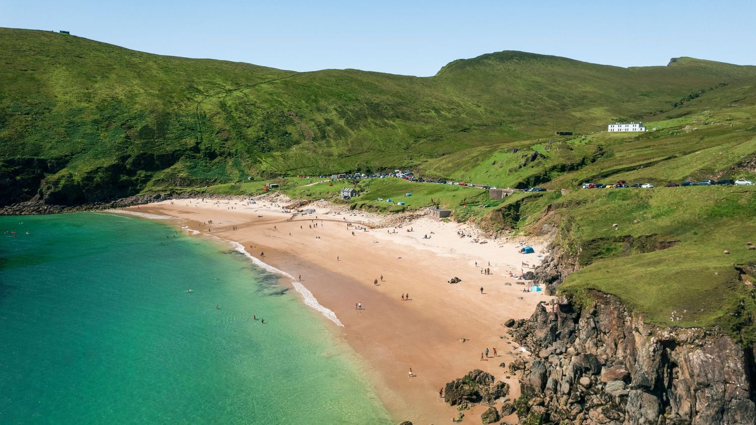 Aerial view of a sandy beach with turquoise water nestled between dramatic green Irish cliffs.