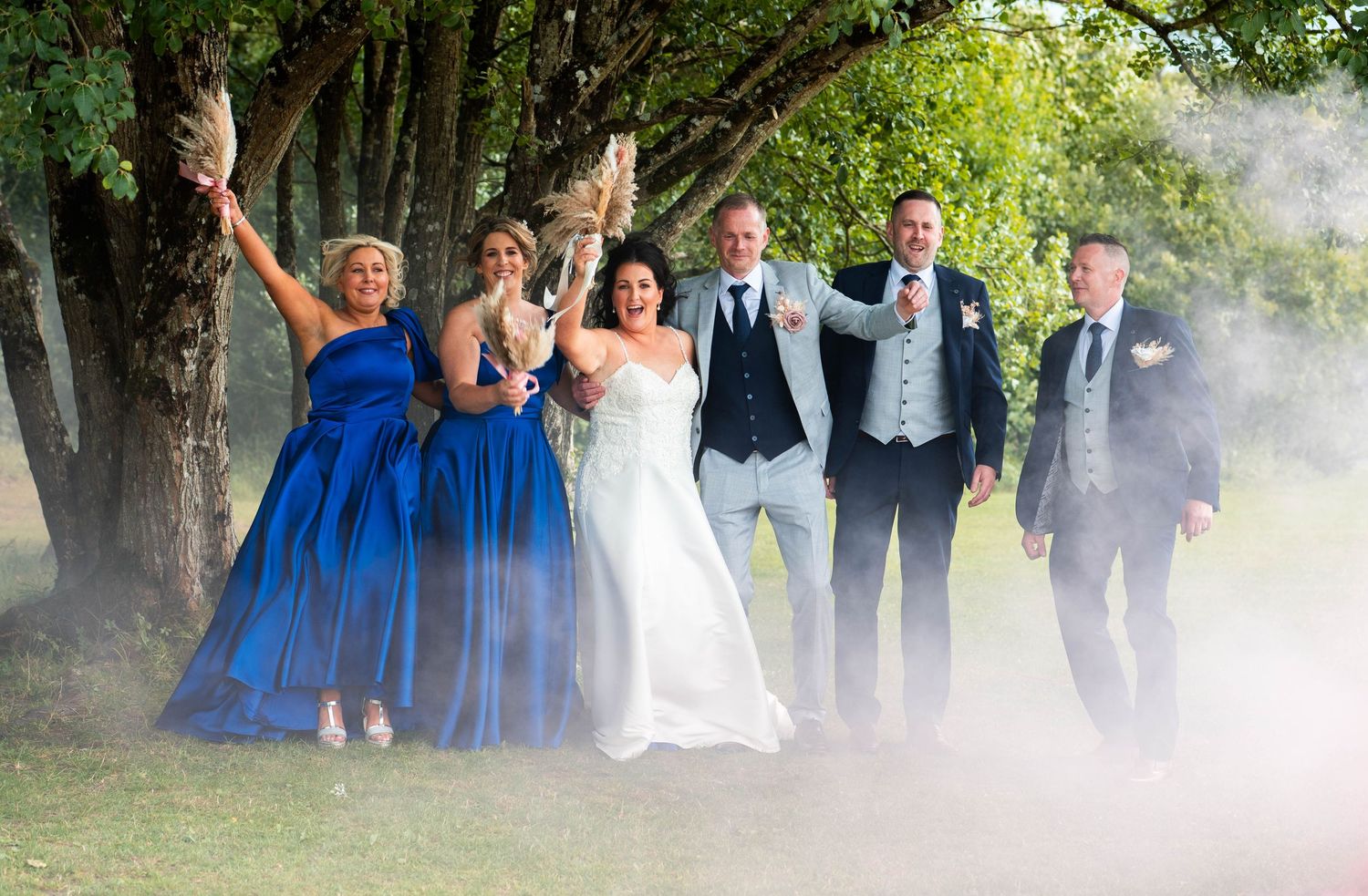 Joyful wedding party celebrates outdoors among trees with dramatic smoke effect, bridesmaids in blue and groomsmen in navy.