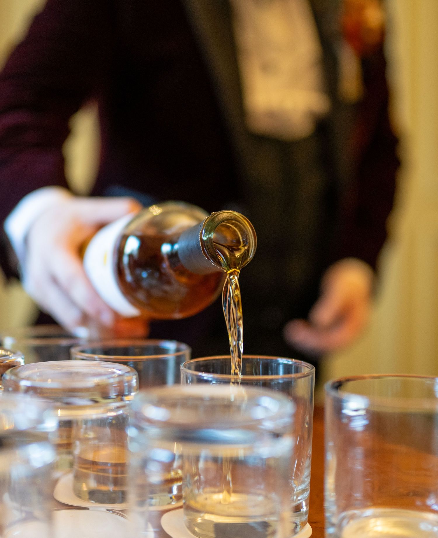 Liquid being poured from a bottle into multiple glasses on a wooden table.