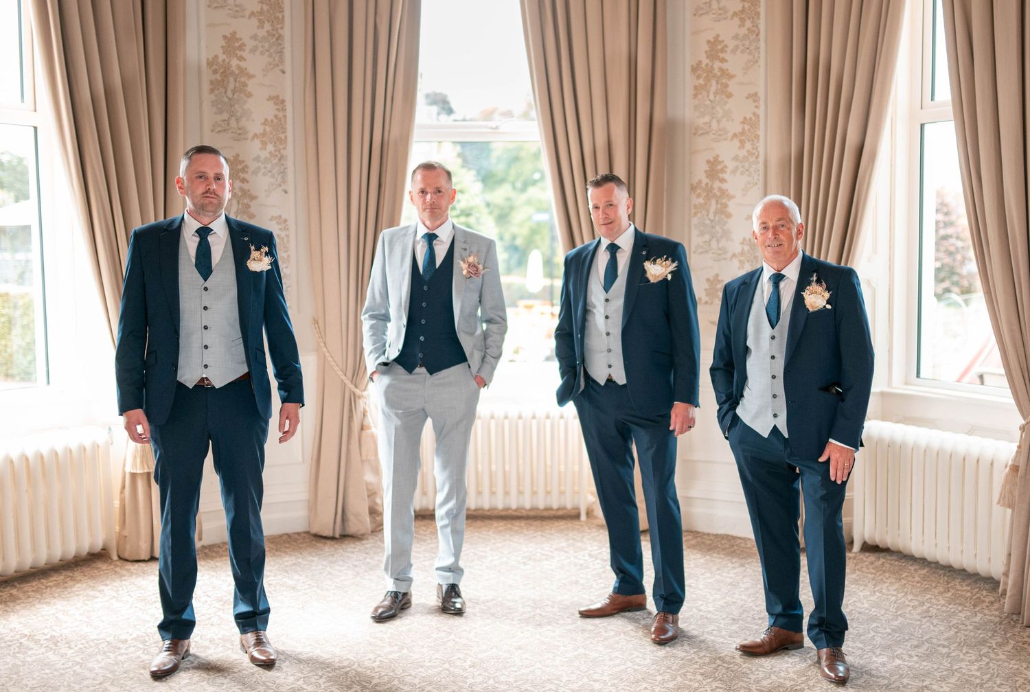 Four groomsmen in navy and grey suits posing elegantly in a bright, curtained wedding venue room.