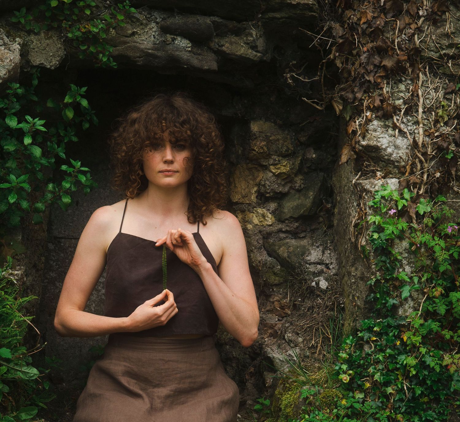 Young woman in brown linen dress holding a stem in a mossy stone archway with ivy.