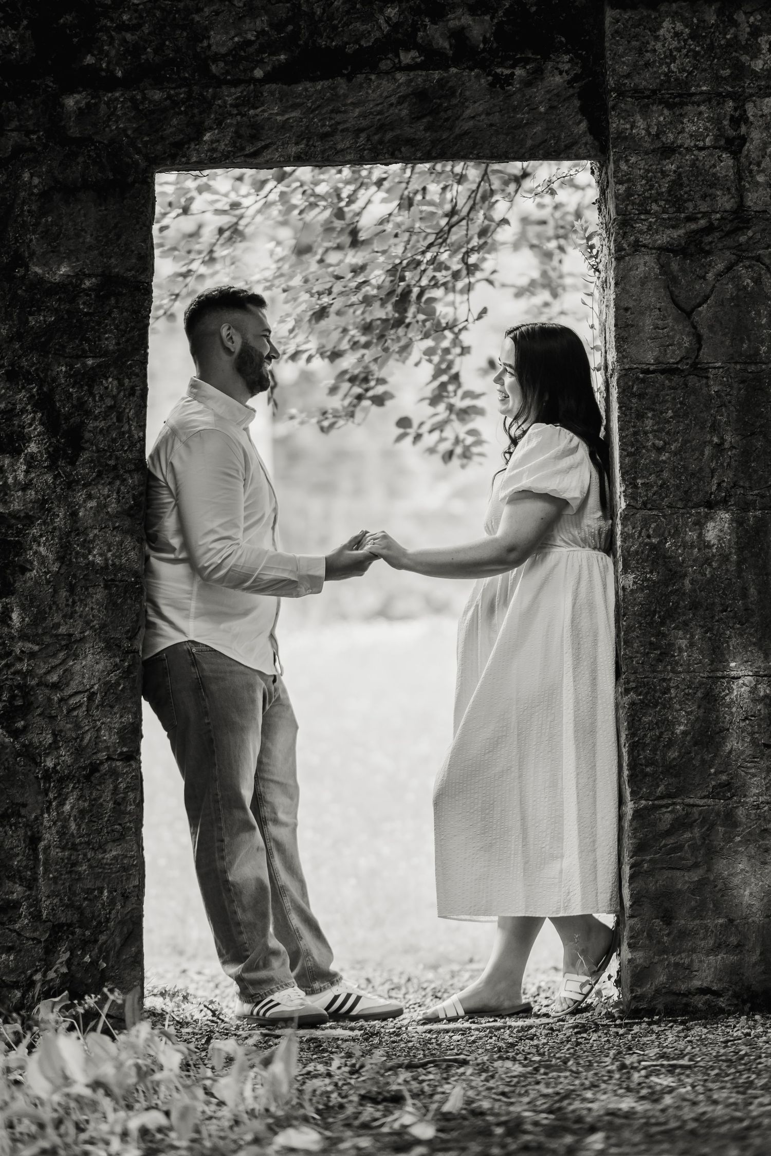 Black and white photo of a couple holding hands in a stone archway framed by trees outdoors.