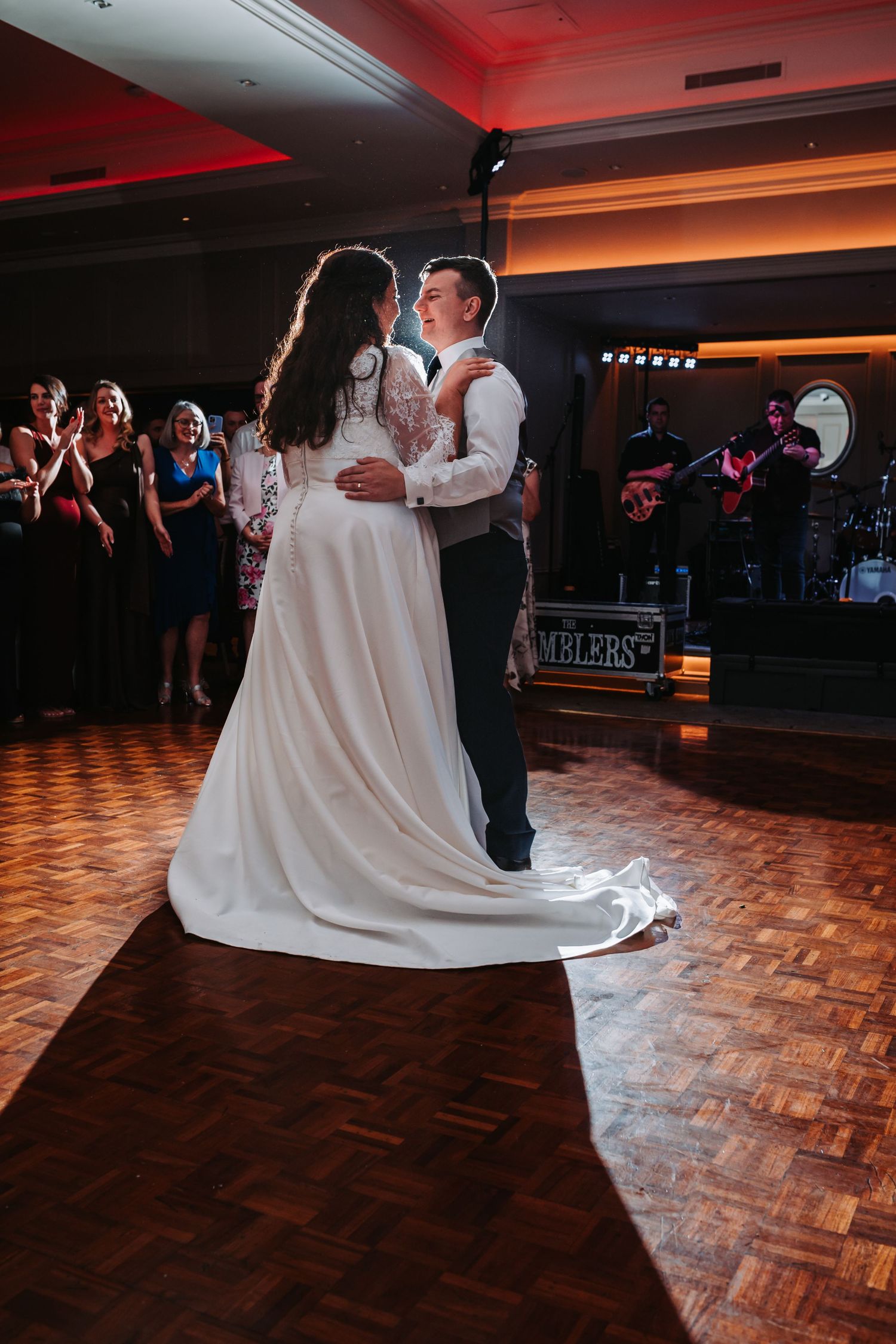Bride and groom share romantic first dance on wooden dance floor with live band and guests watching.