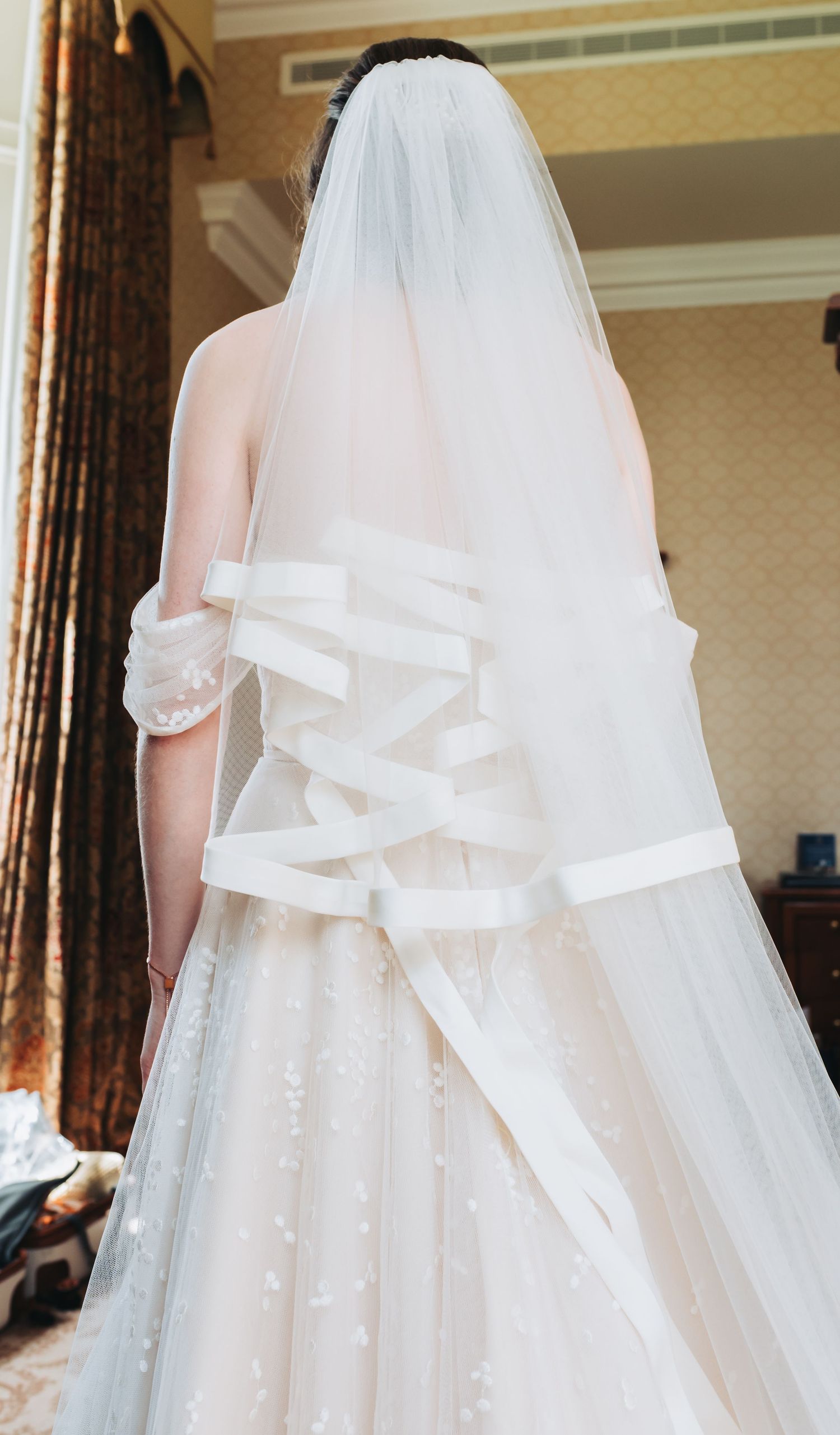 Bride in white lace wedding gown with ribbon corset back and layered veil, viewed from behind.