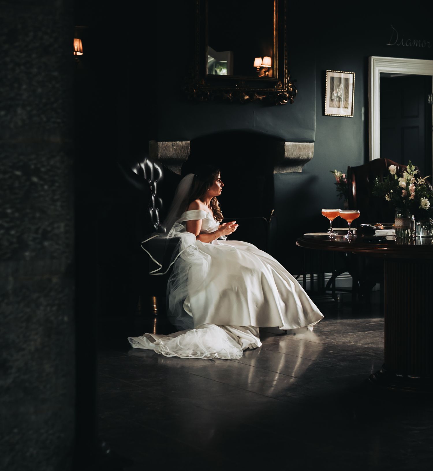 A person in a white wedding dress sits elegantly in a dark leather armchair in a moody room.