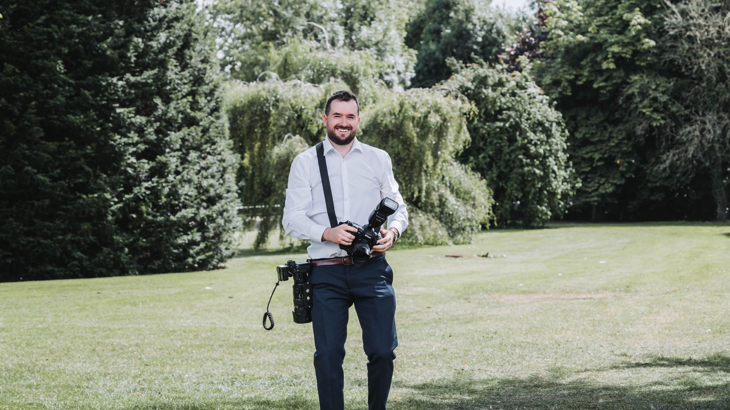 A wedding photographer walks across a lush green lawn during an outdoor summer celebration.