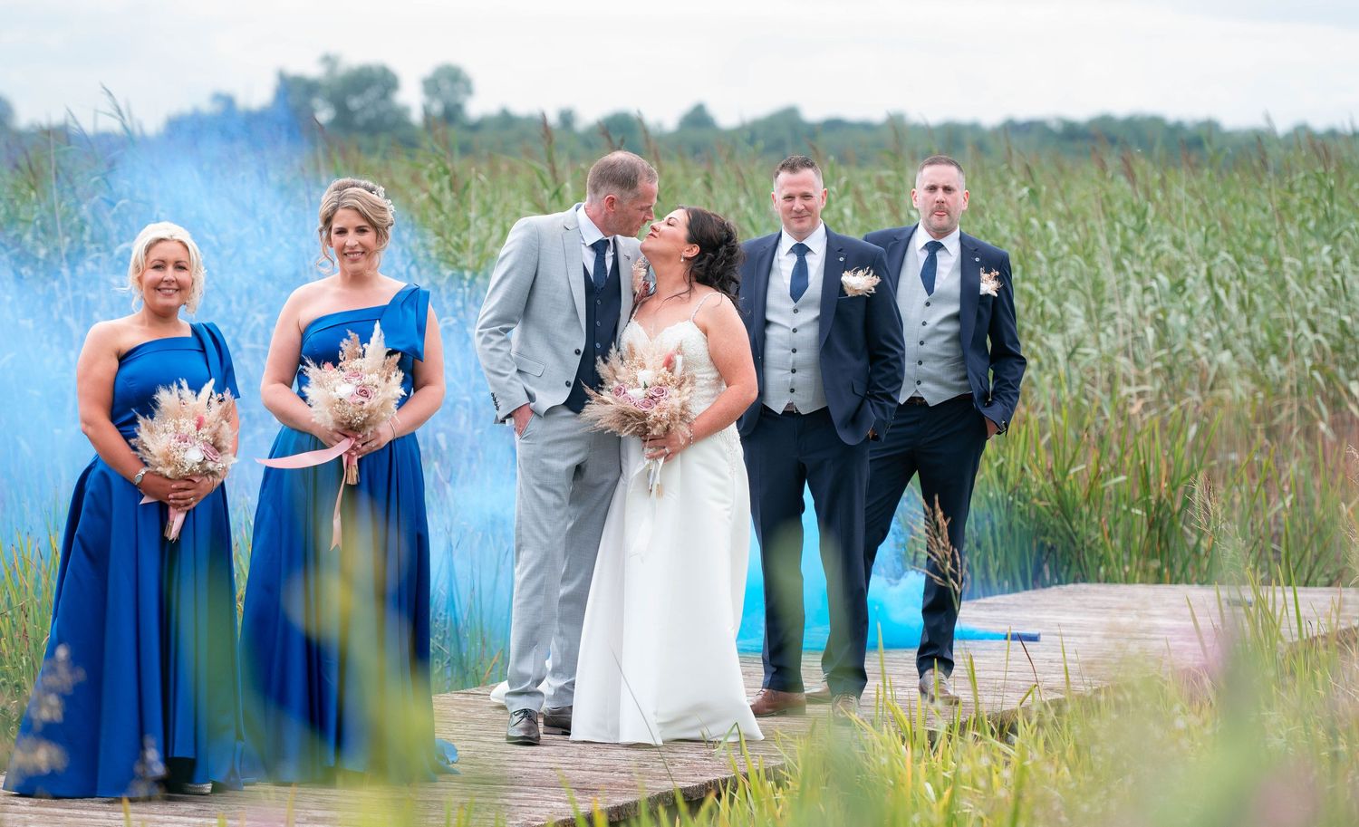 Wedding party poses on a wooden boardwalk surrounded by tall grass and blue sky.