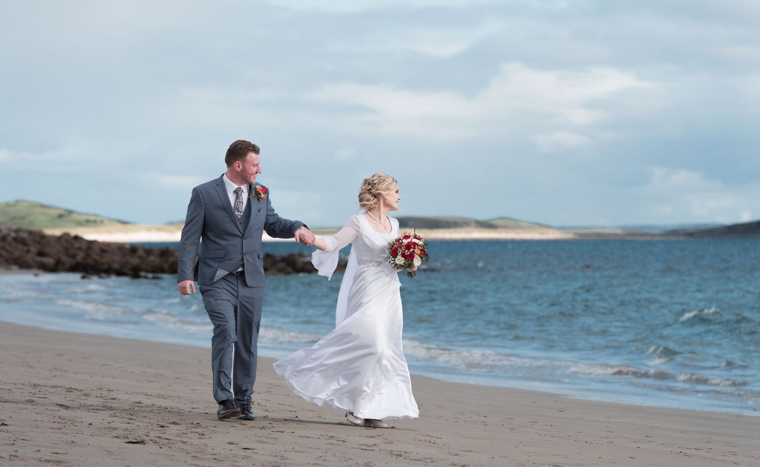 Newlyweds stroll hand in hand along a scenic beach with ocean views.