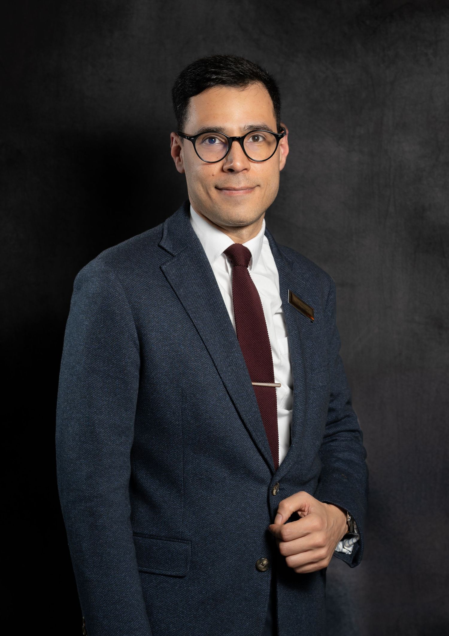 Professional man in blue suit, burgundy tie, and glasses posing confidently for a formal portrait photo.