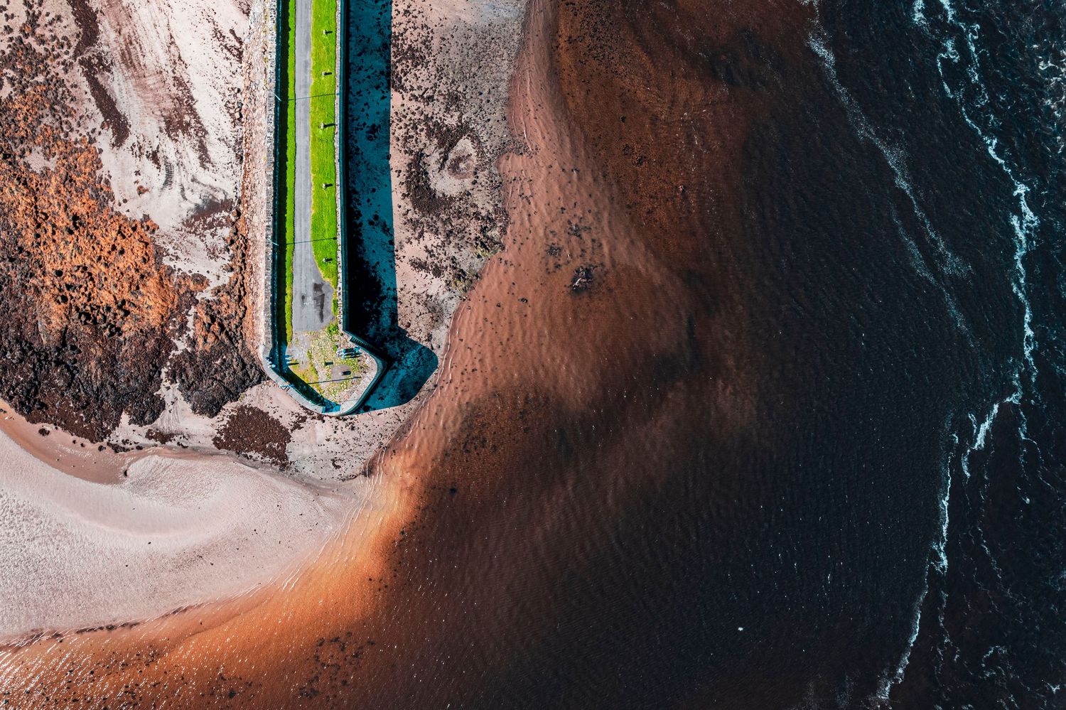 Aerial view of a green pier extending into brown sandy shoreline meeting dark ocean waves.
