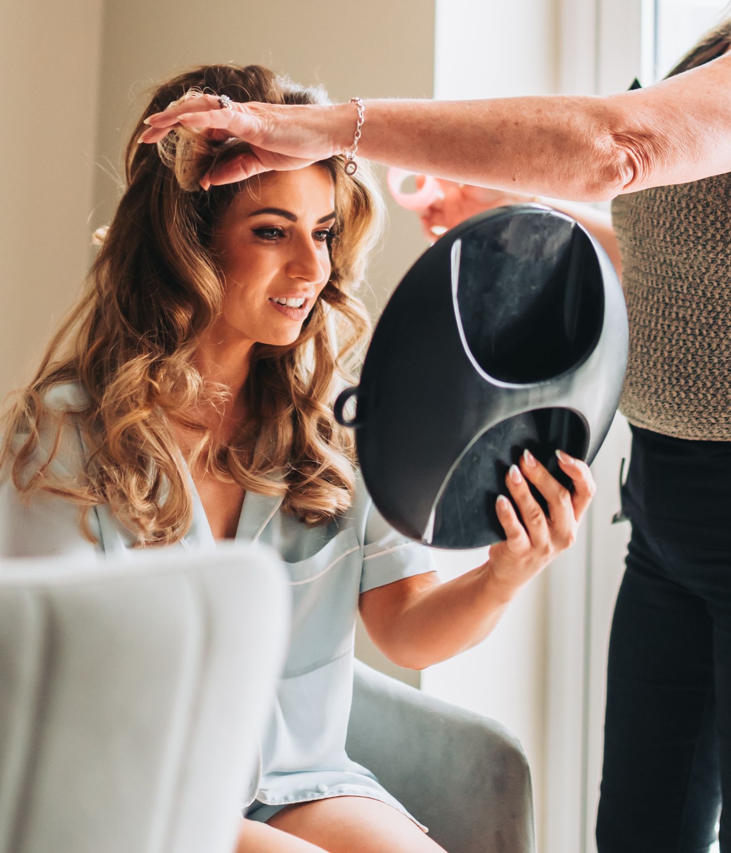 A bride smiles while getting her hair styled, holding a mirror to check her wedding day look.
