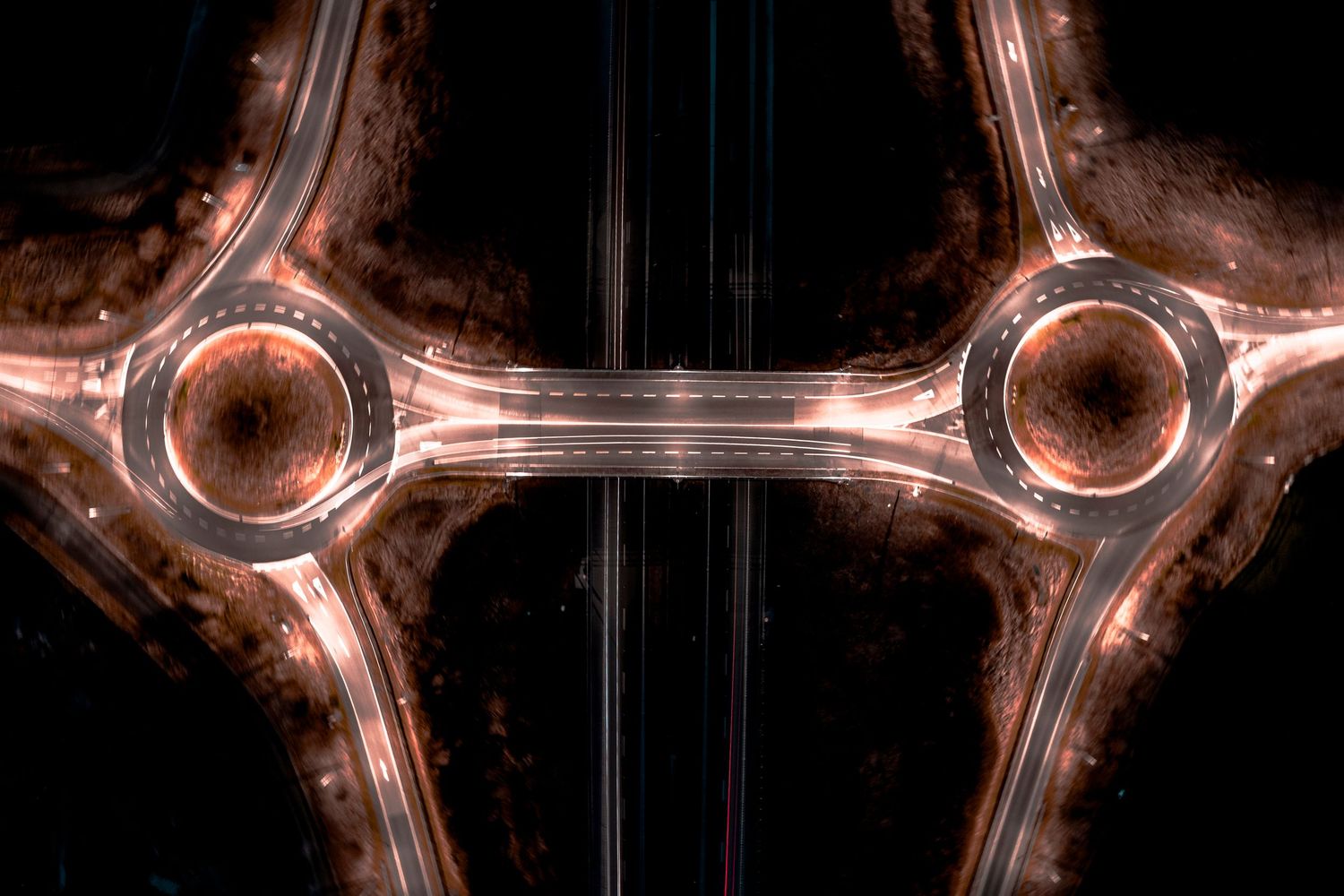 Aerial top-down view of two illuminated roundabouts connected by a highway at night creating a symmetrical pattern.
