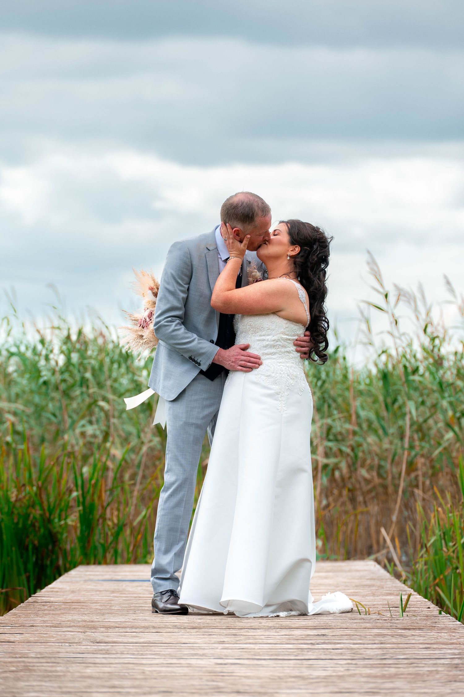 Bride and groom kissing on wooden dock surrounded by tall reeds on cloudy day.