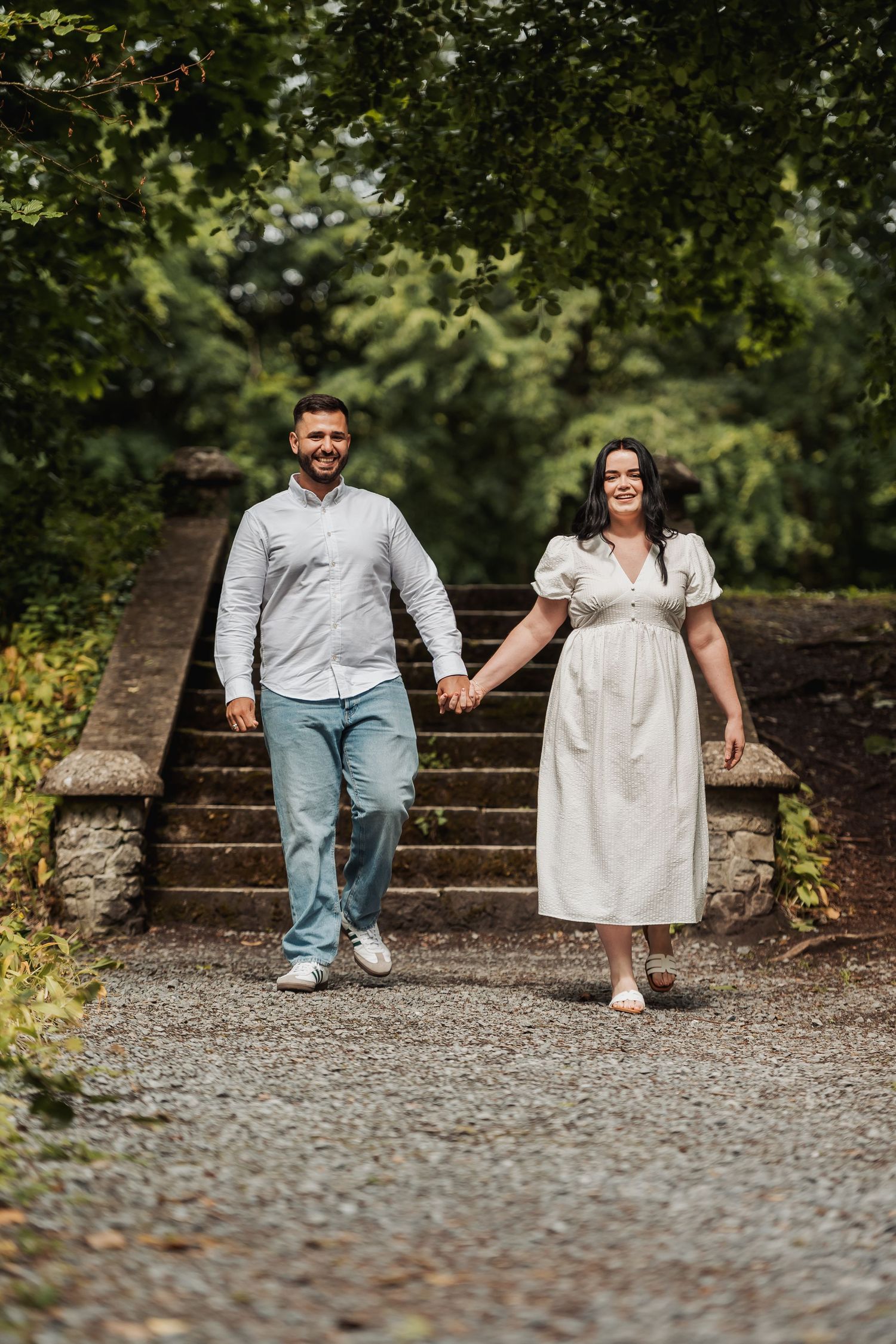 A smiling couple holds hands walking down stone steps surrounded by lush green trees in an outdoor garden setting.