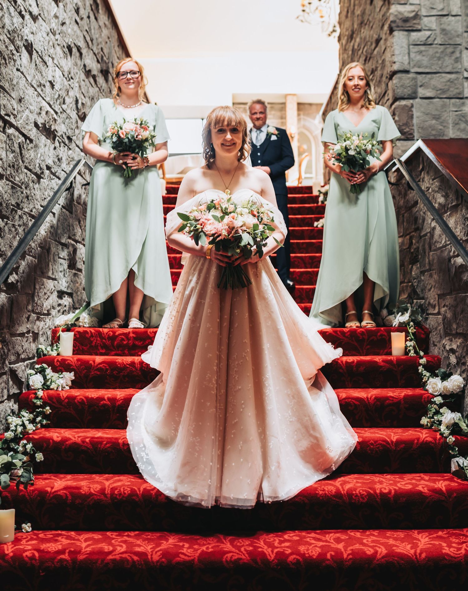 Bride in blush ballgown holds bouquet on red carpeted stairs flanked by mint green bridesmaids at elegant wedding venue.