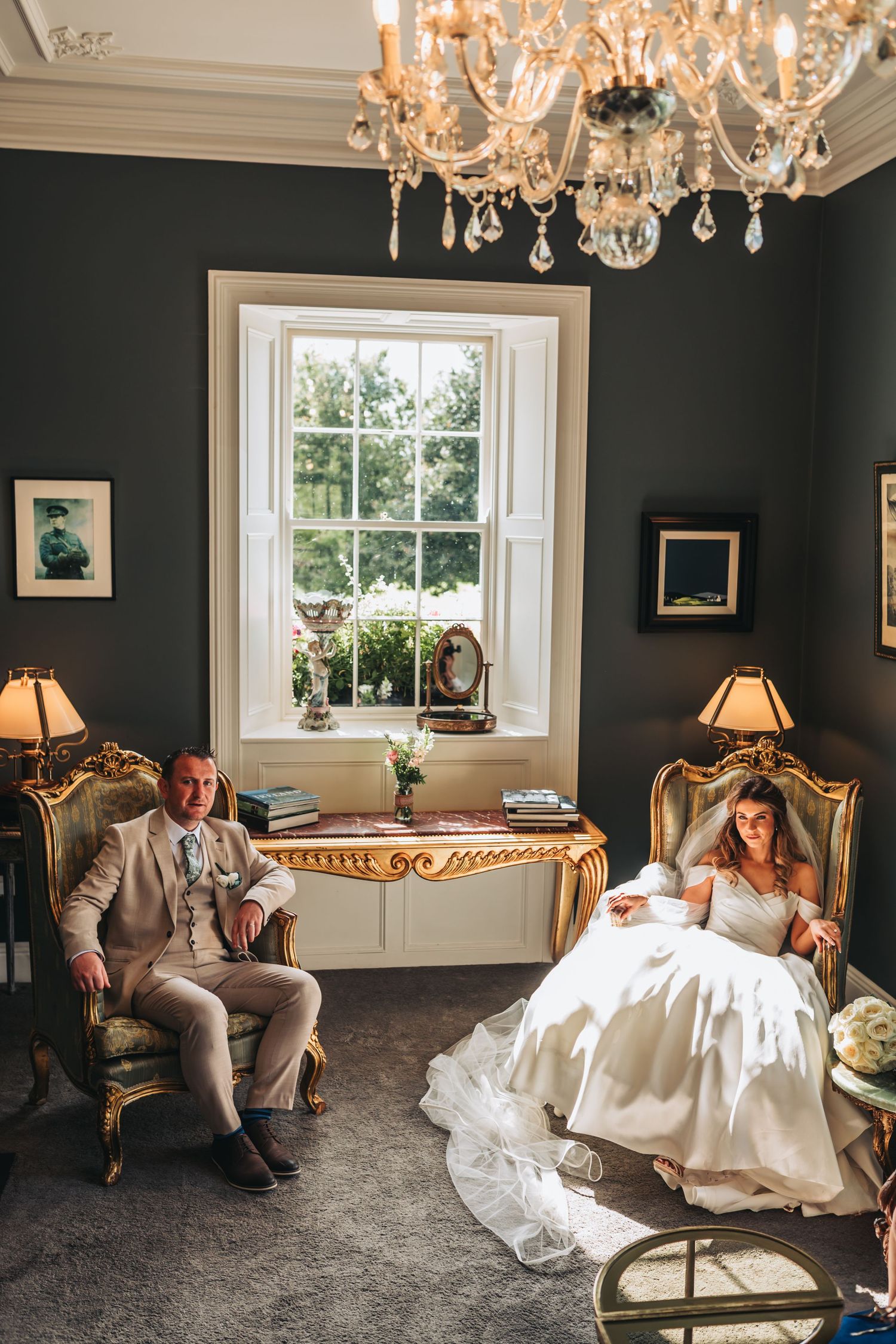 Elegant wedding couple lounges in a dark room with crystal chandeliers and ornate furniture.
