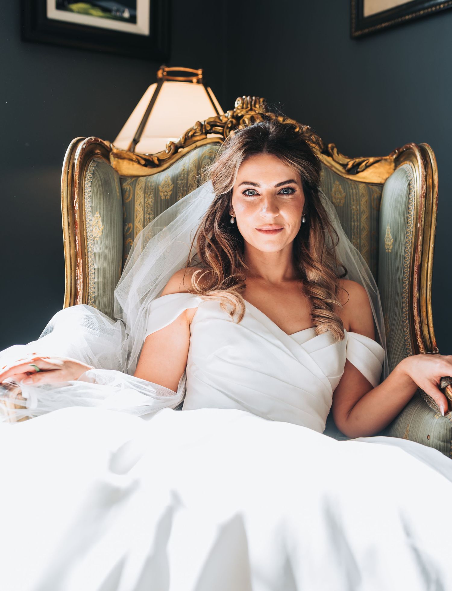 Bride in white wedding dress and veil lounges elegantly on vintage chair during bridal boudoir photoshoot.