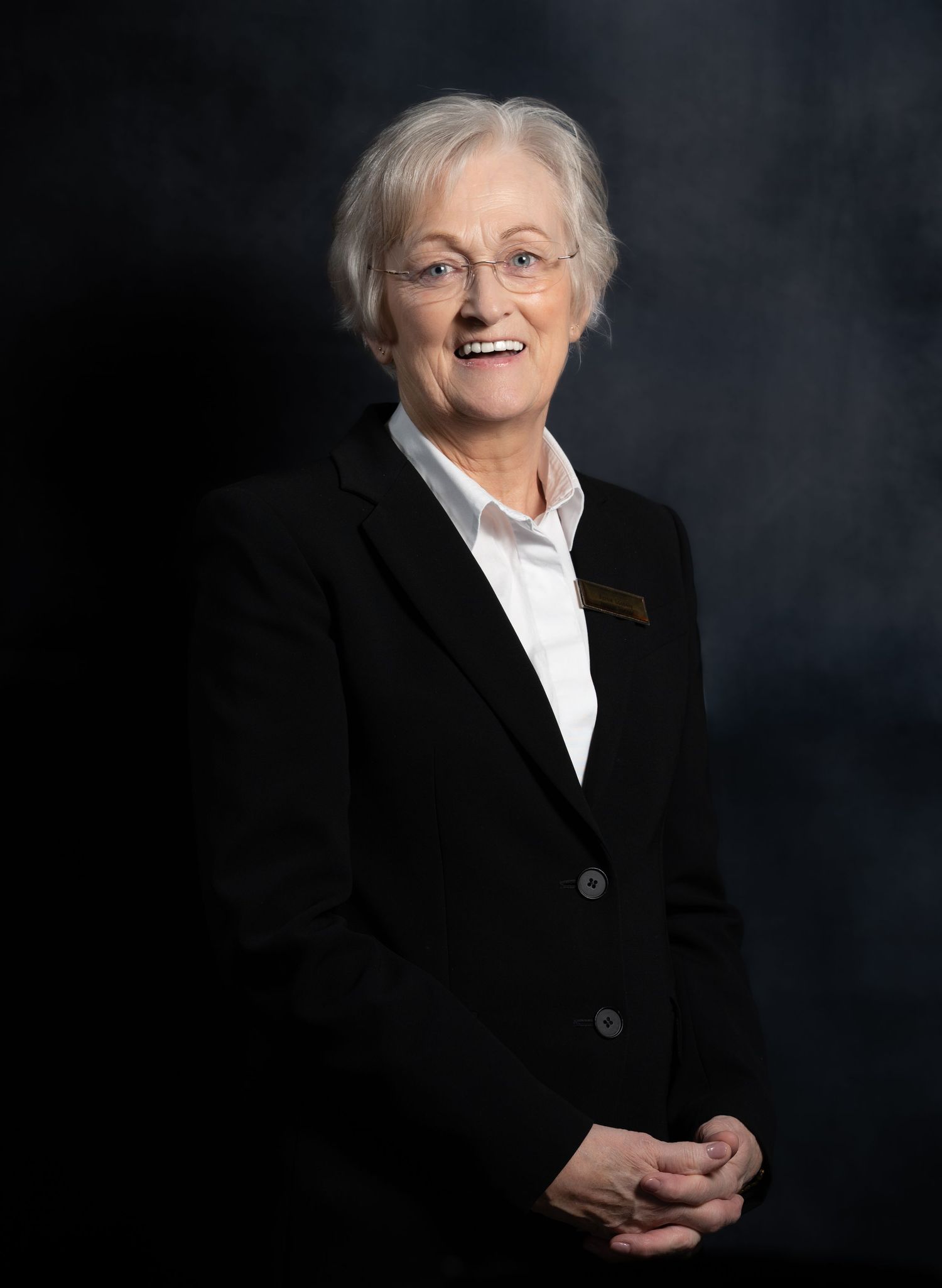Smiling elderly woman with gray hair and glasses wearing a black blazer and white shirt poses for a professional portrait.