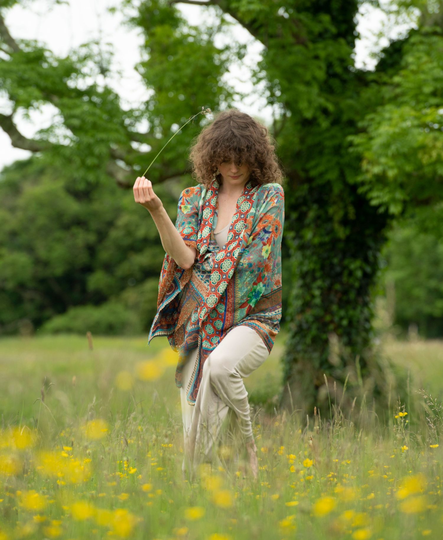 Woman in colorful boho kimono holding a wire in a wildflower meadow with trees in background.