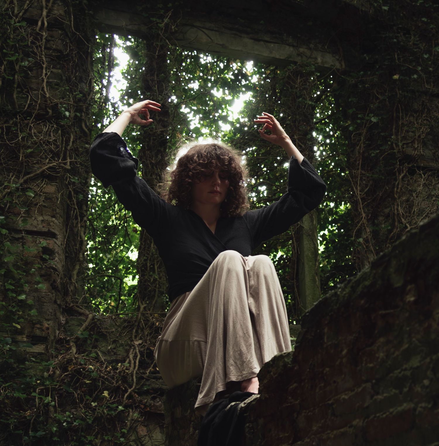 Woman with curly hair sitting in stone ruins surrounded by lush green ivy and foliage.