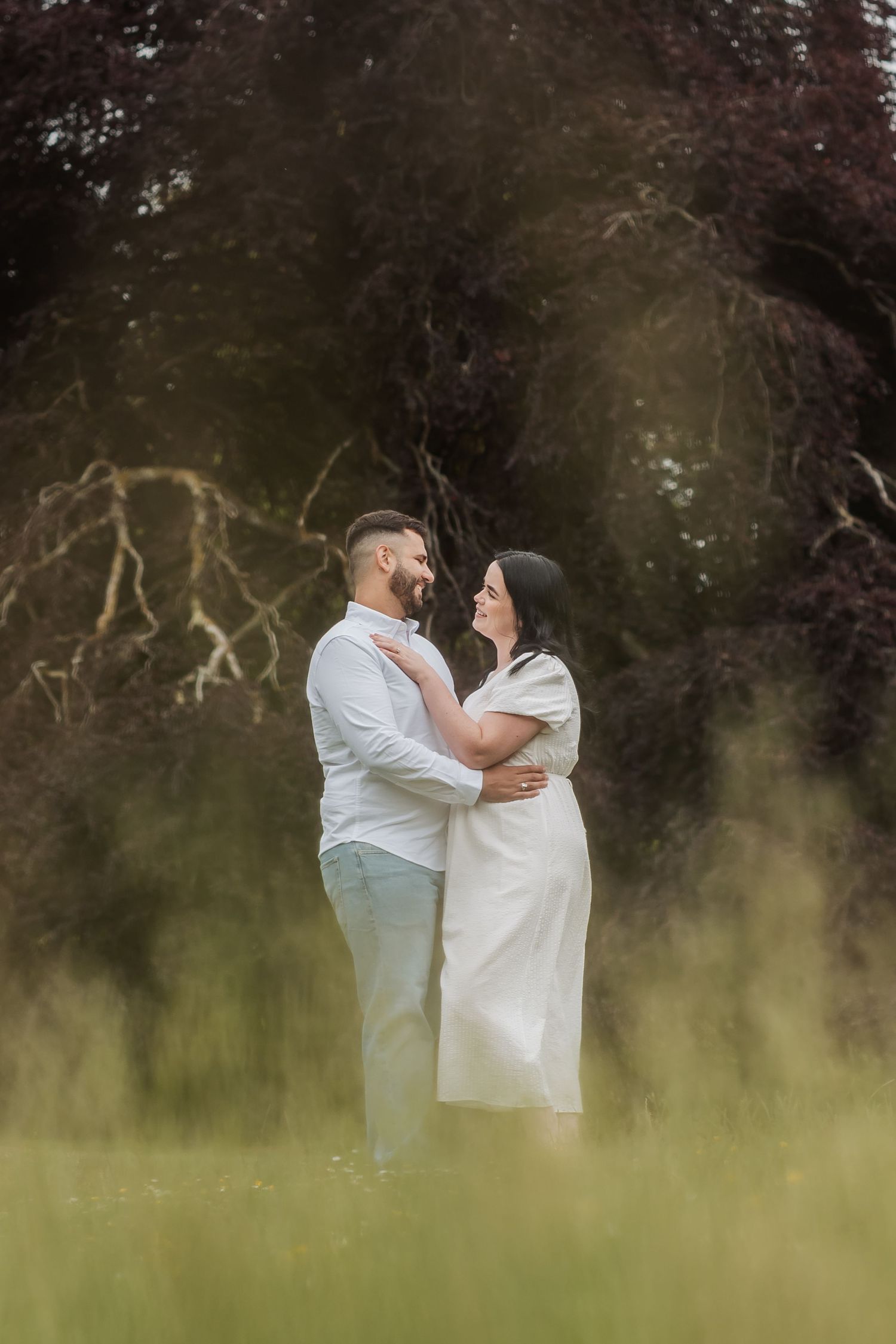 Couple embracing and gazing at each other in a lush green field with dark trees in the background.