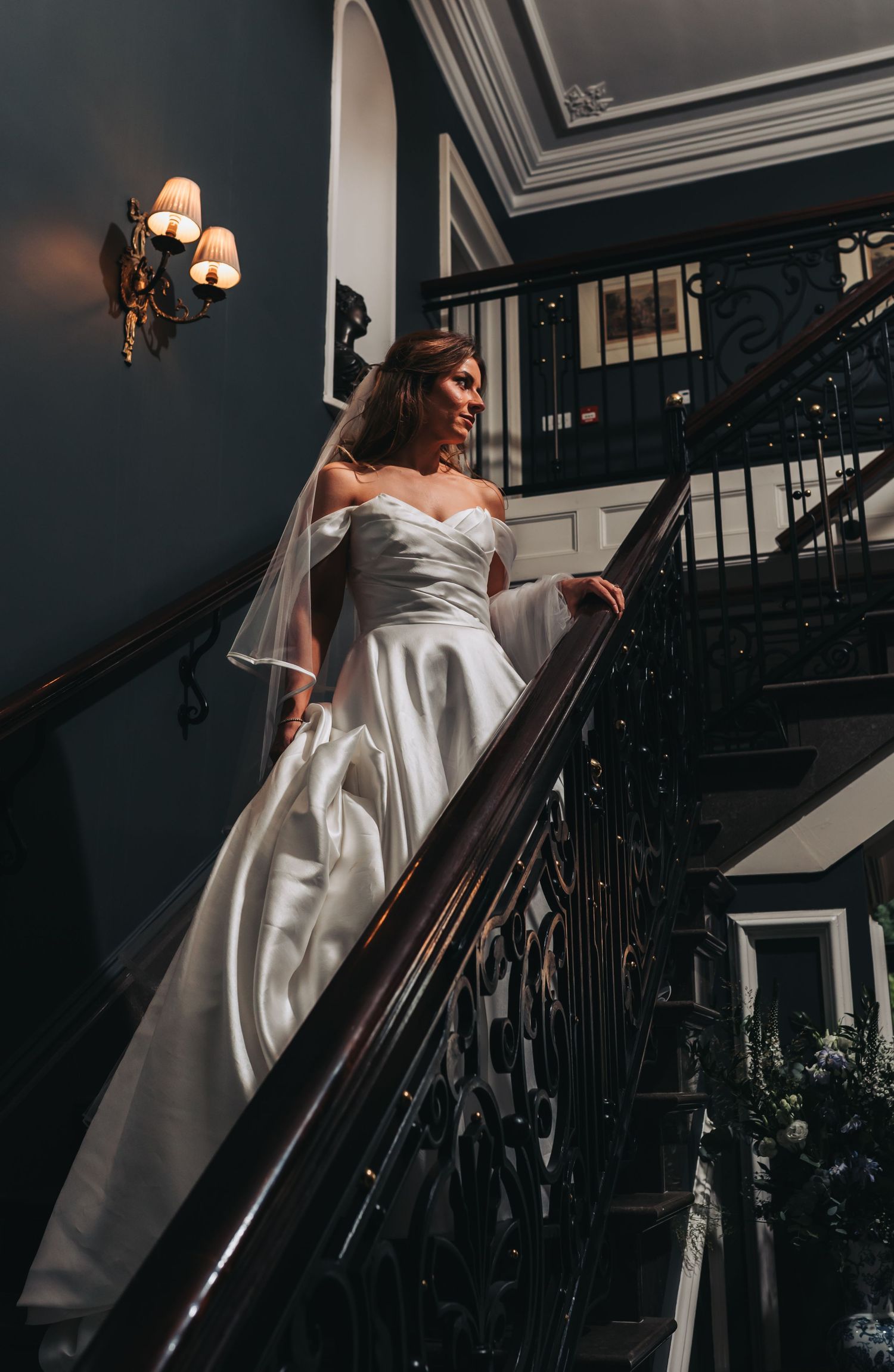 Elegant bride in white satin gown holding railing on grand staircase in dramatic lighting.
