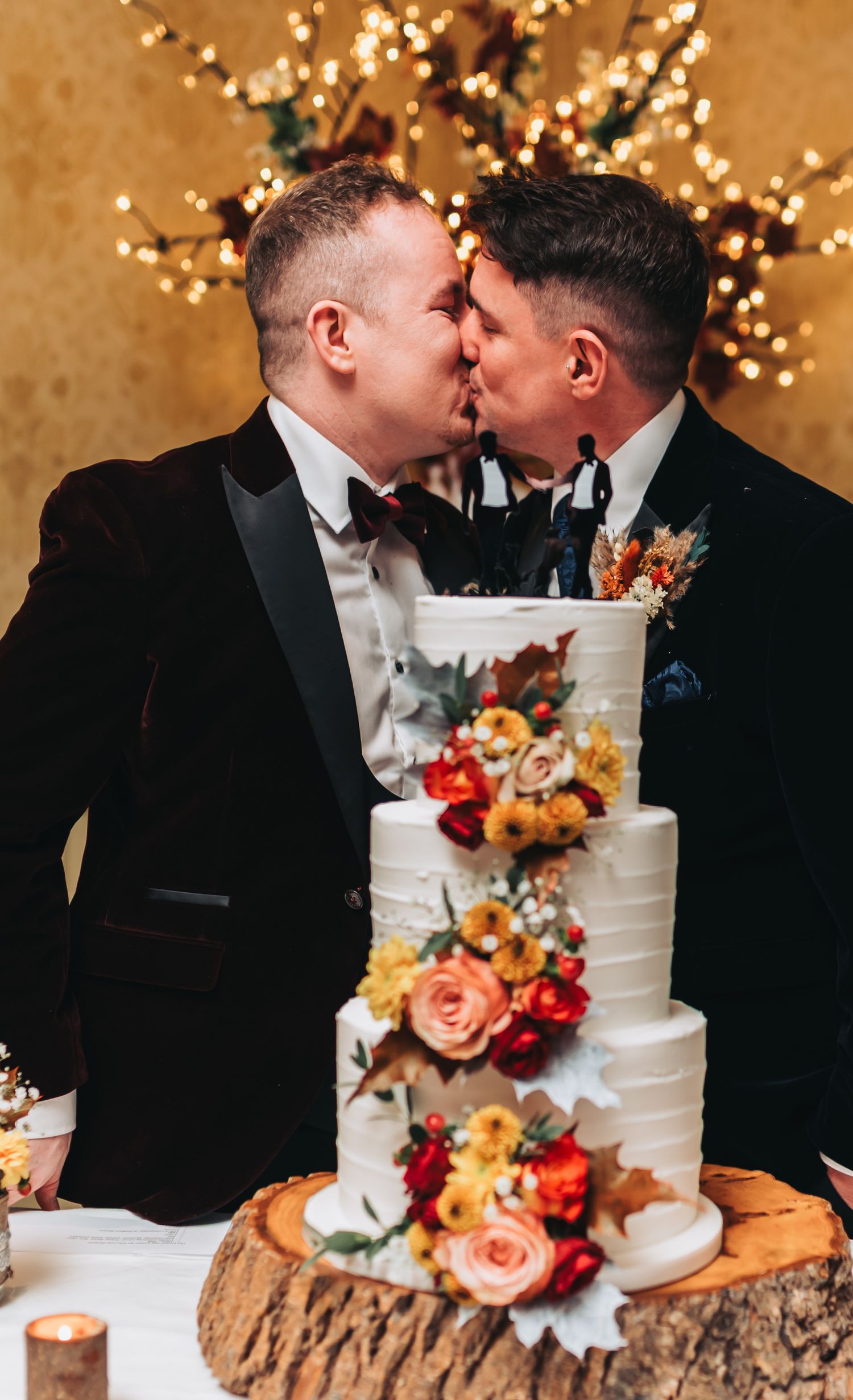 Wedding cake decorated with fall flowers on a wooden stand with twinkling lights in background.