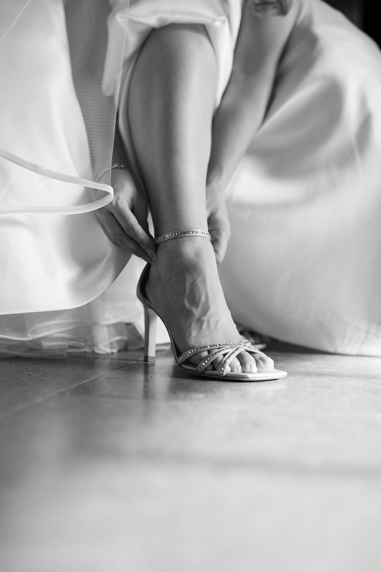 Black and white close-up of bride fastening strappy heeled wedding shoes with anklet.