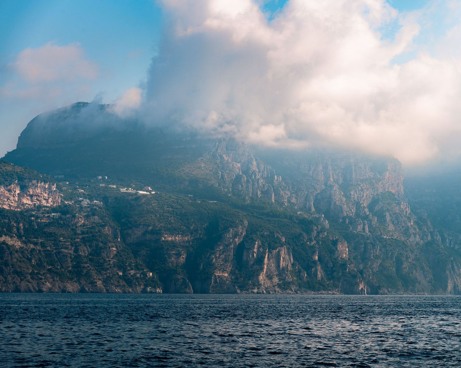 Dramatic rocky Amalfi Coast cliffs rise from choppy blue sea beneath cloudy skies.