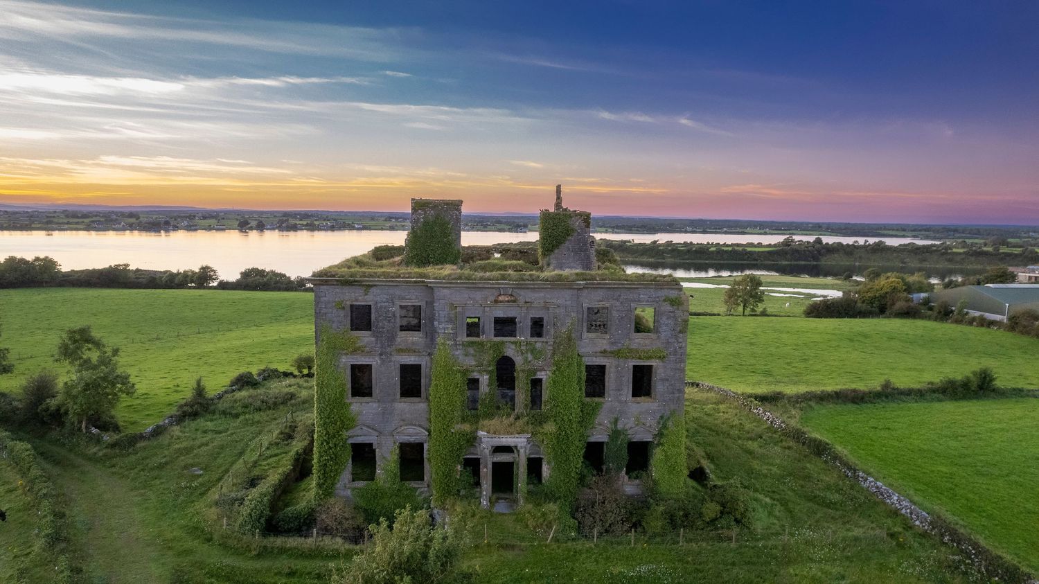 Aerial view of an abandoned ivy-covered mansion ruin at sunset in lush Irish countryside.