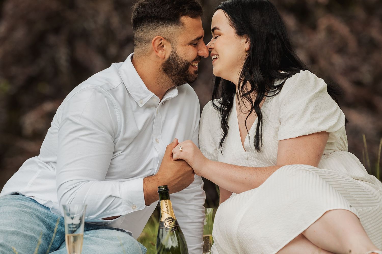 Romantic couple in white outfits sharing an intimate moment with champagne bottle during an outdoor engagement photo session.
