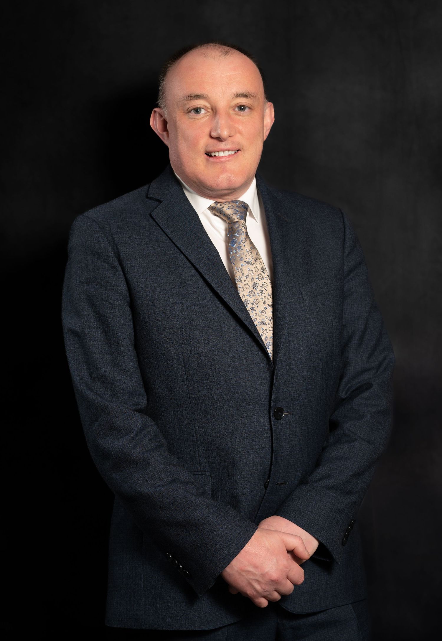 Professional man in dark suit and patterned tie poses for formal business portrait against dark background.