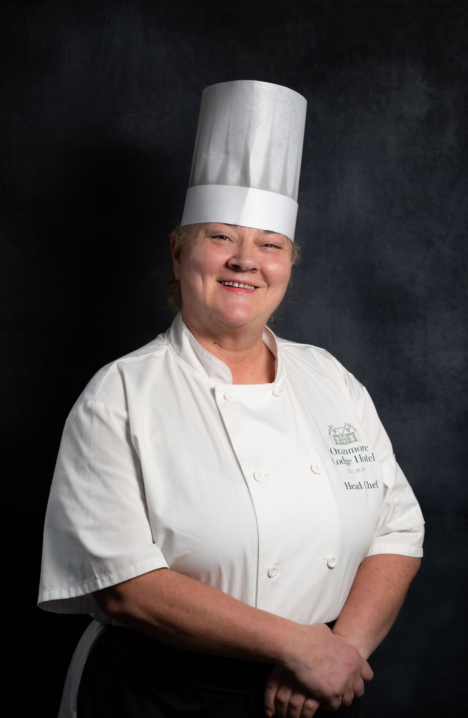 Smiling female head chef in white uniform and tall toque hat poses with arms crossed at restaurant.