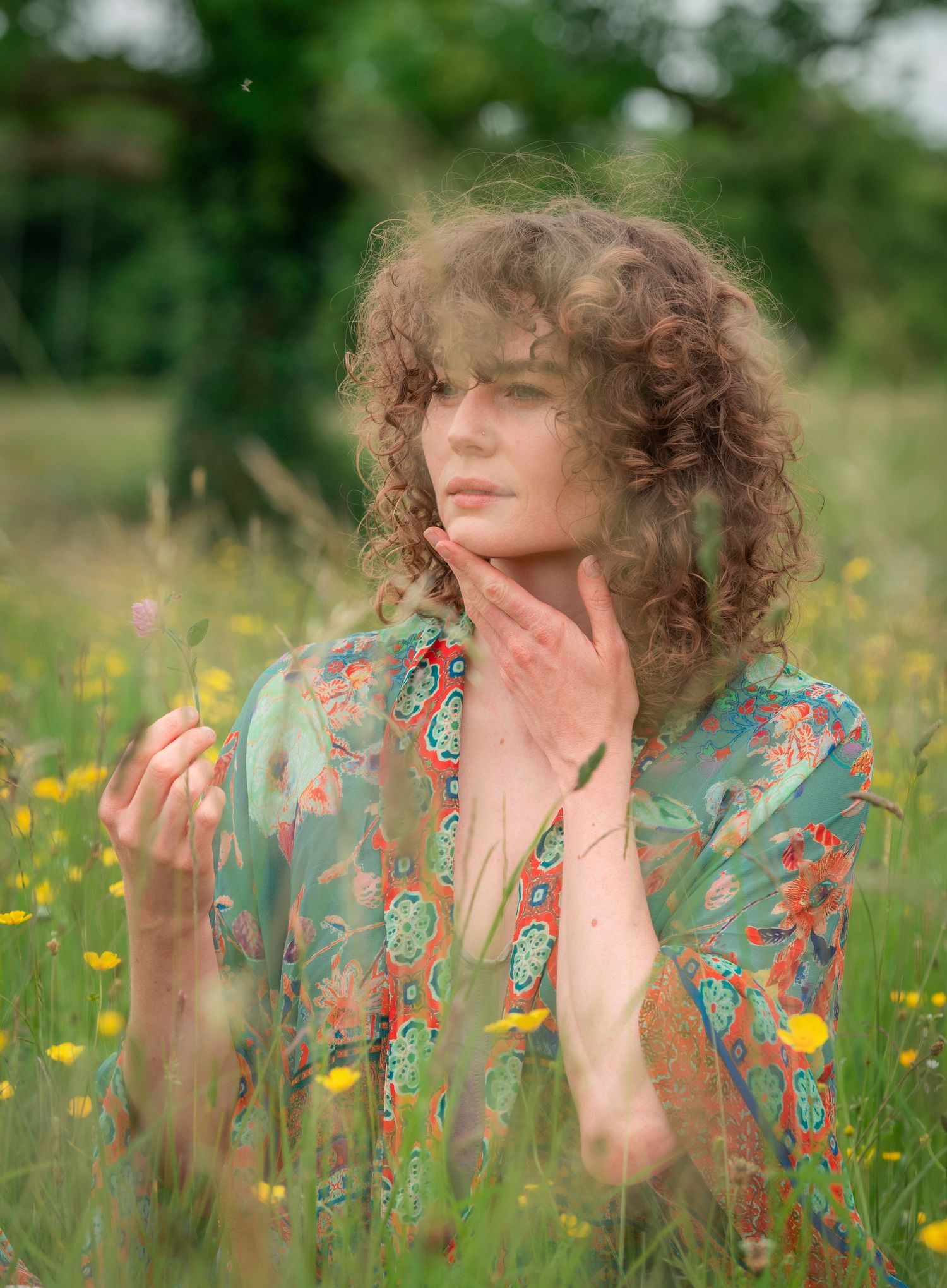 Young woman with curly hair in floral dress sitting in wildflower meadow.