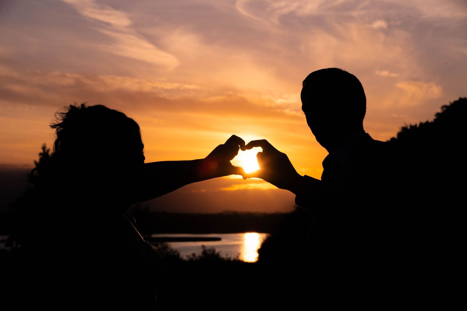Couple silhouetted against golden sunset forming heart shape with hands near lake.