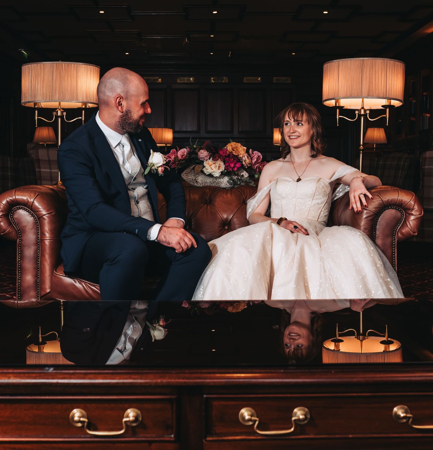 Bride and groom sit on leather sofa in dark elegant room with warm lamp lighting.