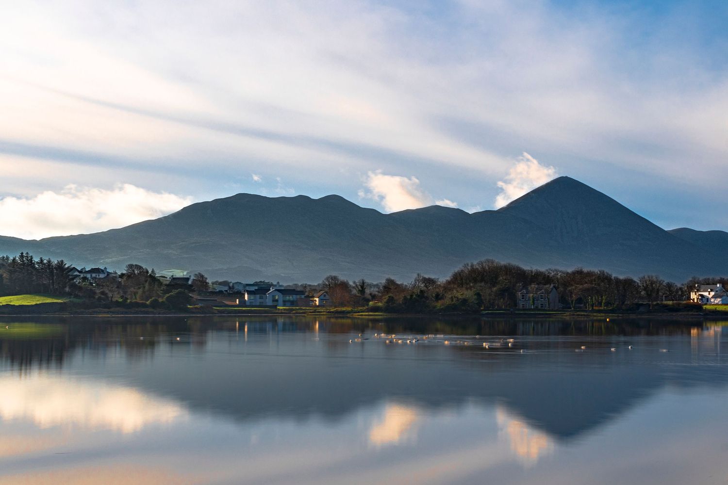 Serene lake reflecting mountains and clouds at dusk, with a small village nestled along the tranquil shoreline.