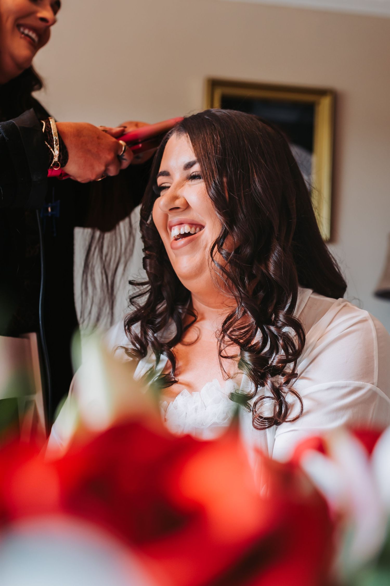 Bride laughing joyfully while getting her dark curly hair styled on her wedding morning.