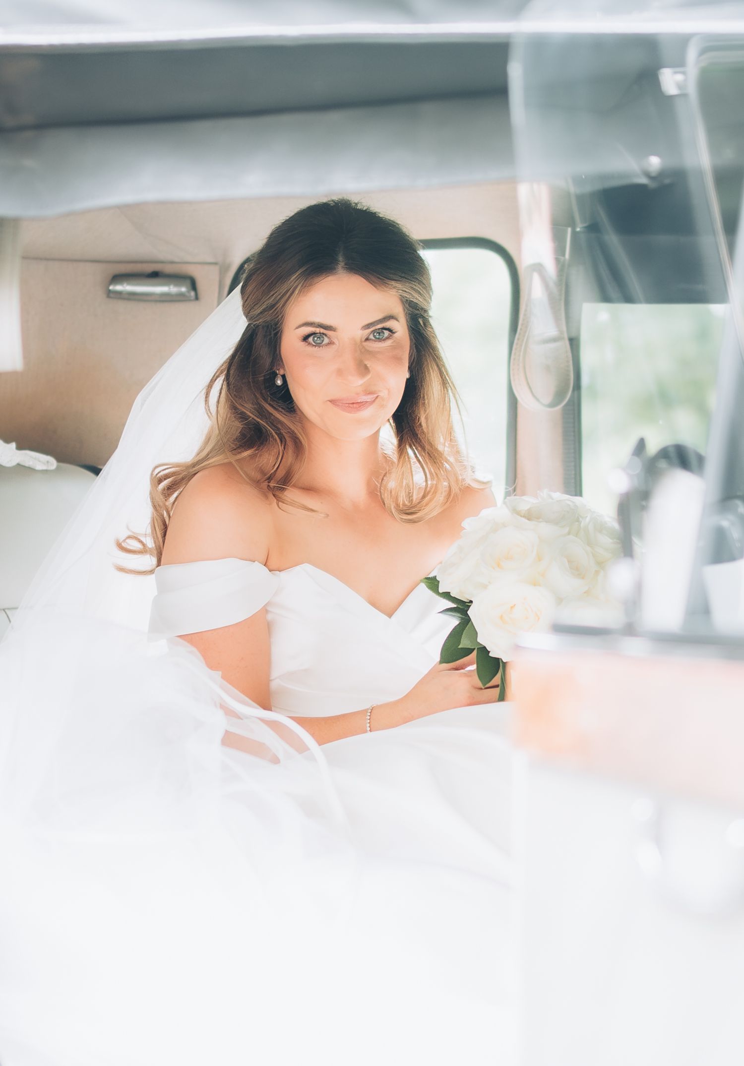 Beautiful bride in off-shoulder gown holding white bouquet sitting in vintage car.