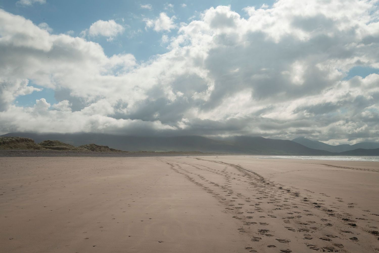 Vast empty sandy beach with footprints leading into the distance under dramatic cloudy skies and distant mountains.