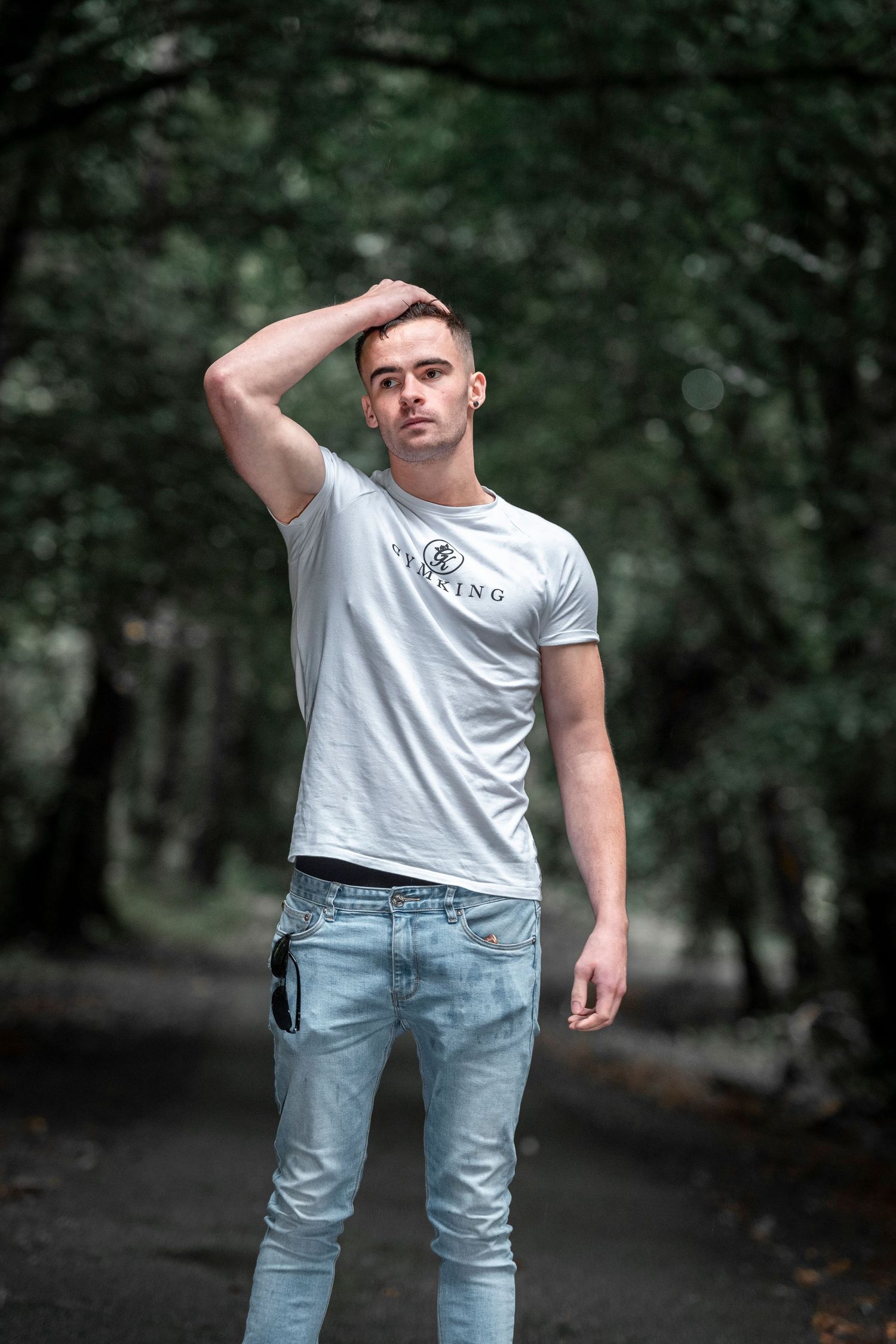 Young man in white t-shirt and jeans posing in green forest path.